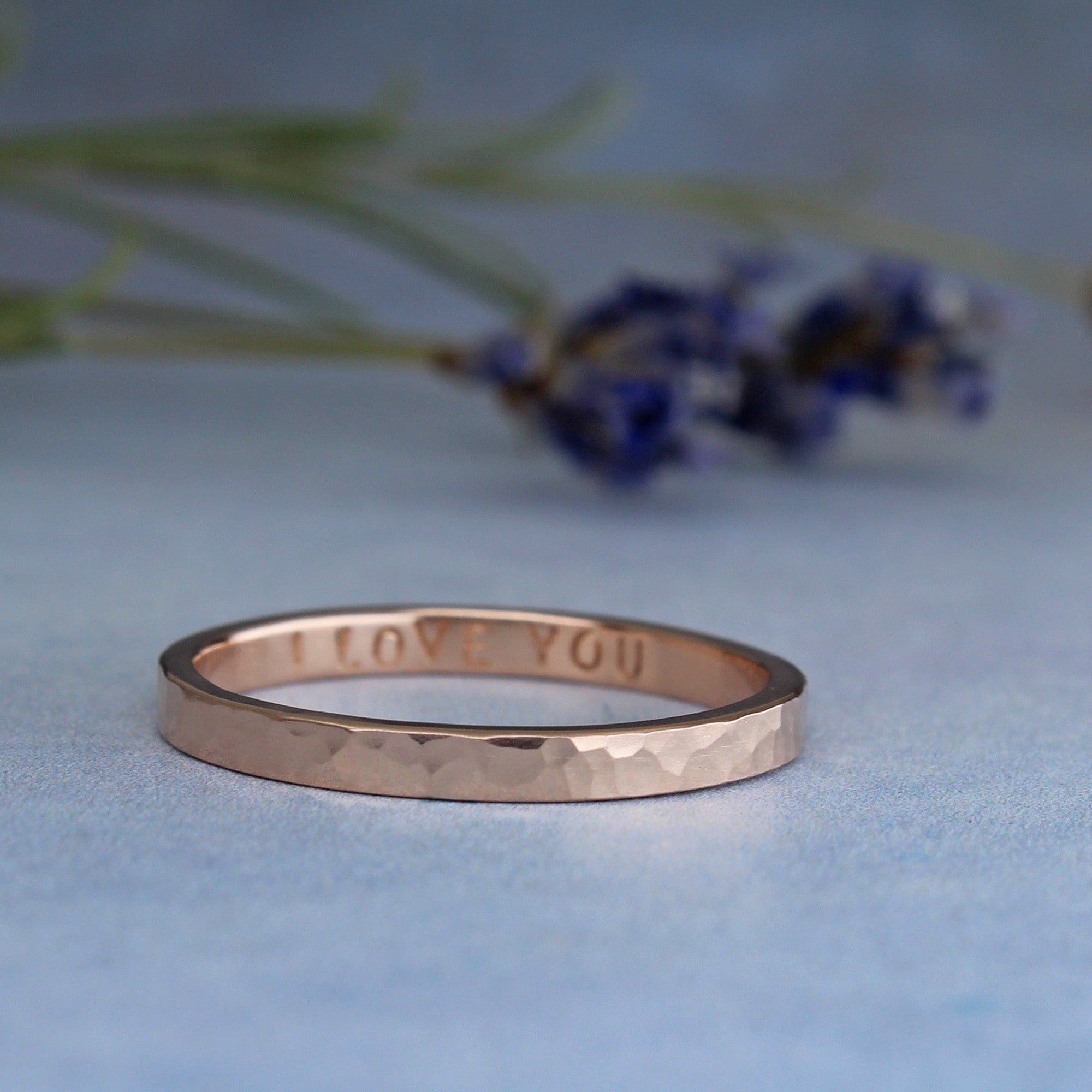Rose gold ring with 'I LOVE YOU' engraving on a light blue background with blurred flowers.