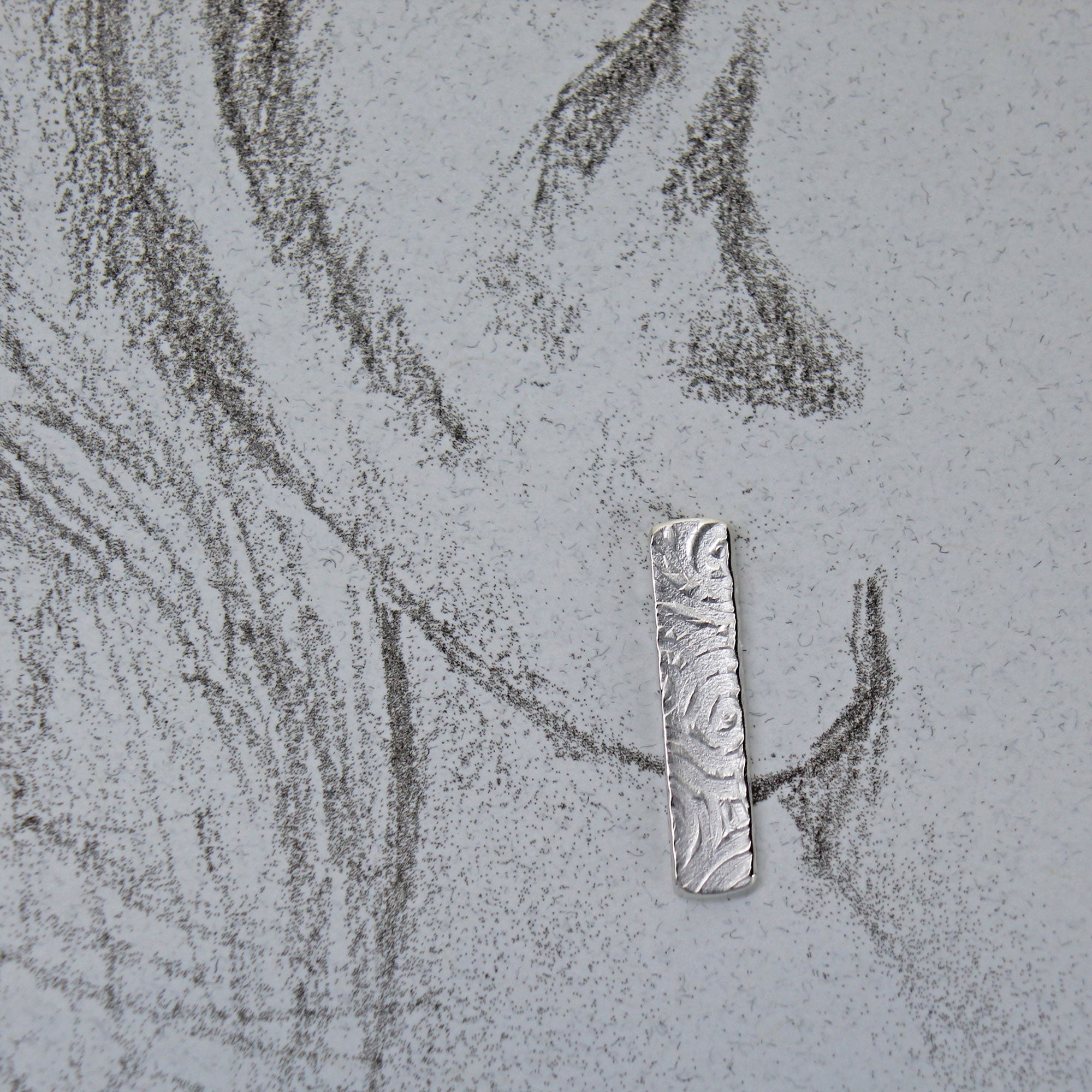 Silver pendant on a textured gray surface with pencil sketch of life size ear