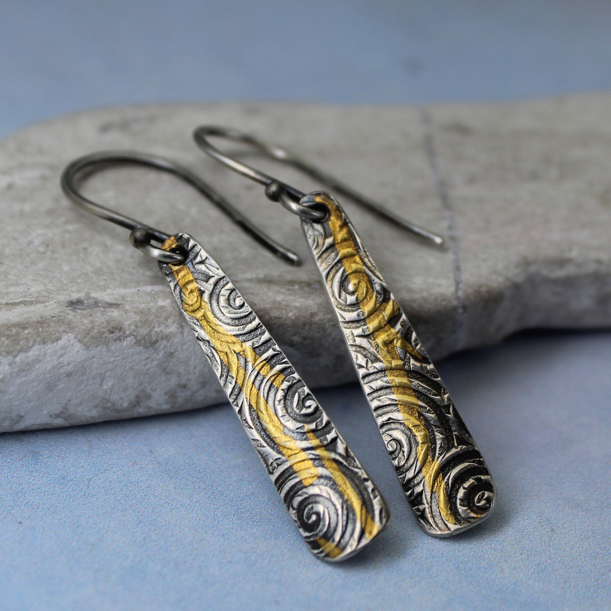 Pair of silver and gold patterned earrings on a stone surface with a blue background