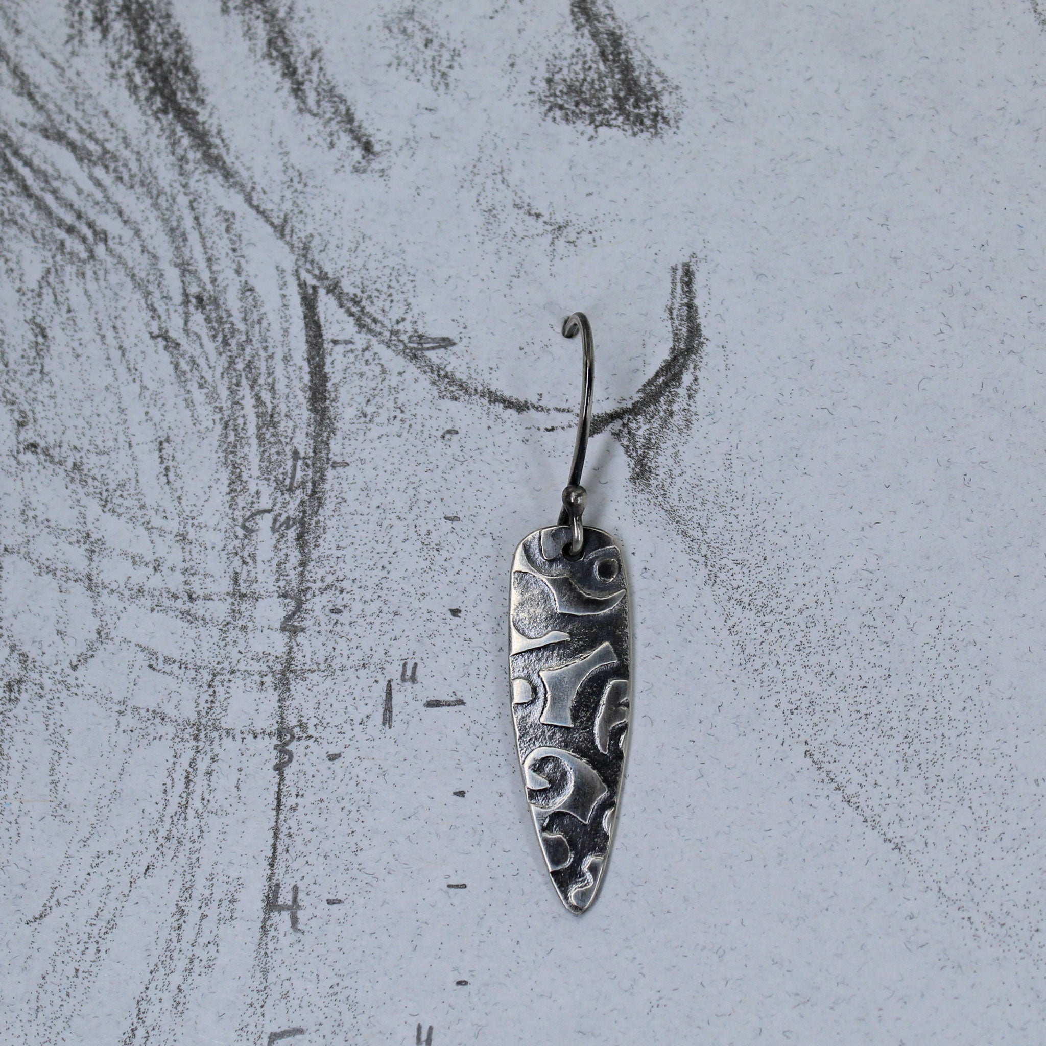 Handmade silver shield dangle earring shown on a pencil drawing of a life size ear.