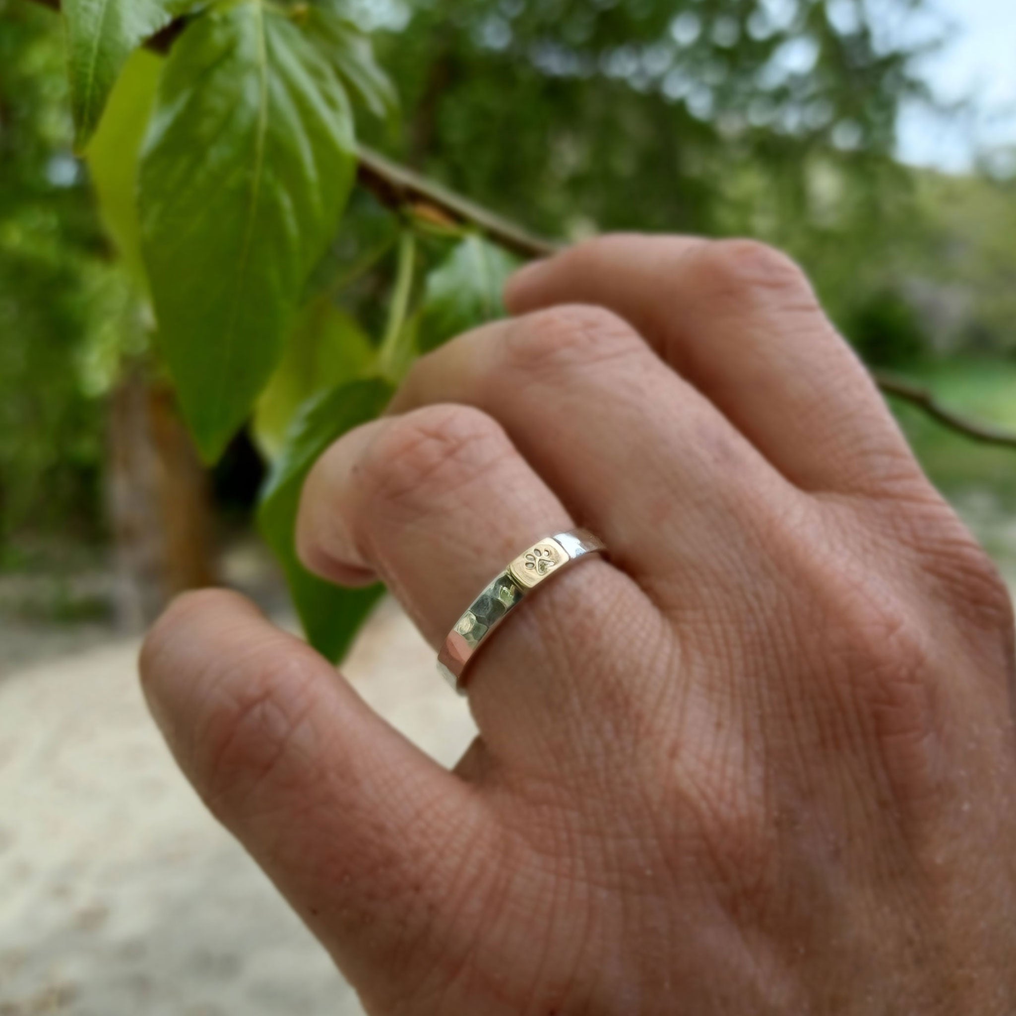 Hand wearing a silver and gold ring with a dog paw symbol, shown with a blurred natural background